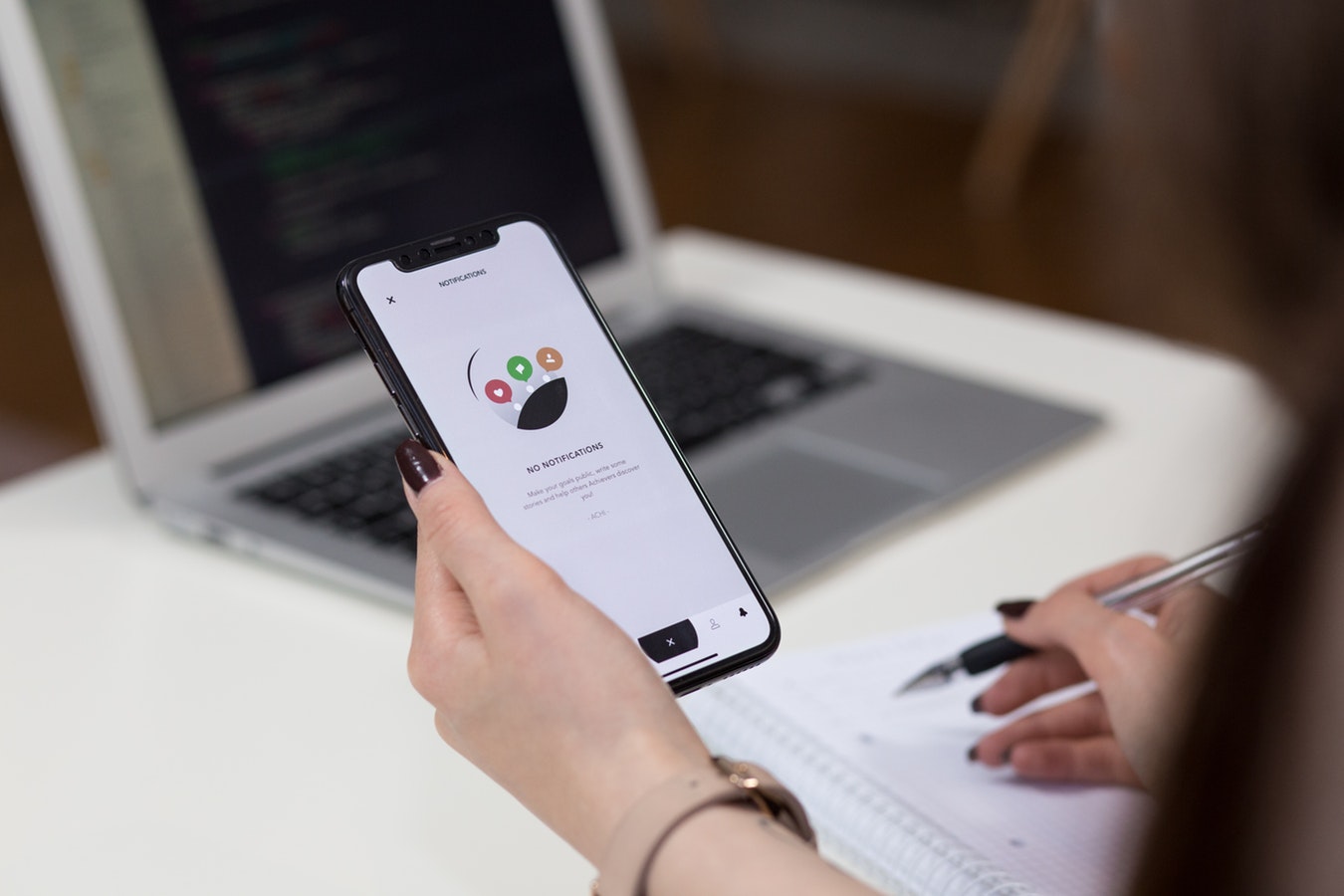 An image of a person writing on a notepad holding an iphone with a laptop in the background
